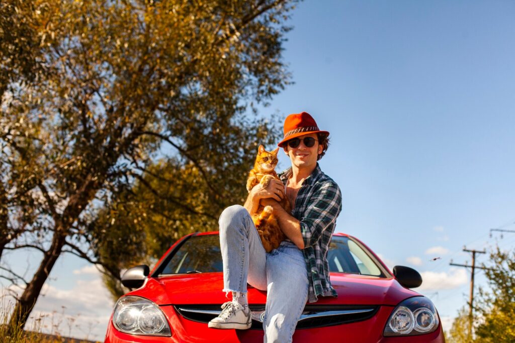 A young man wearing a red hat, sunglasses, and a plaid shirt sits on the hood of a red car, holding a ginger cat, with a scenic outdoor background.